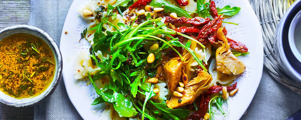 Mediterraner Nudelsalat auf einem Teller bestreut mit Pinienkernen, daneben eine Schale mit Vinaigrette