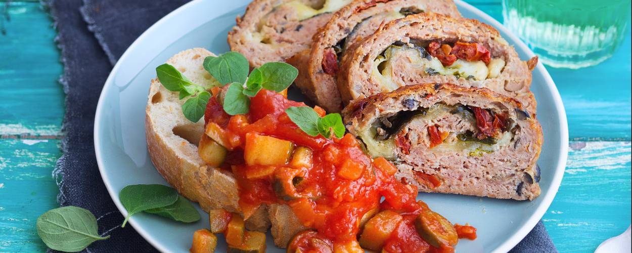Vier Stücke Hackbraten mit Mozzarella-Gemüse-Füllung angerichtet auf einem Teller mit Ciabatta und Zucchini-Tomatensoße