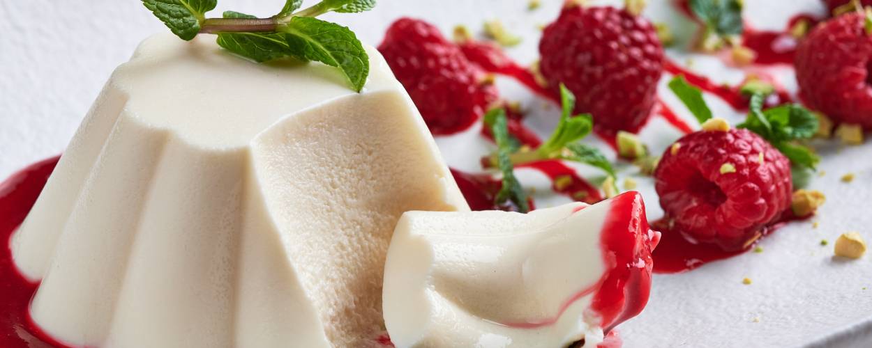 Panna Cotta auf einem Spiegel aus Himbeersoße, daneben frische Himbeeren und Minzblätter