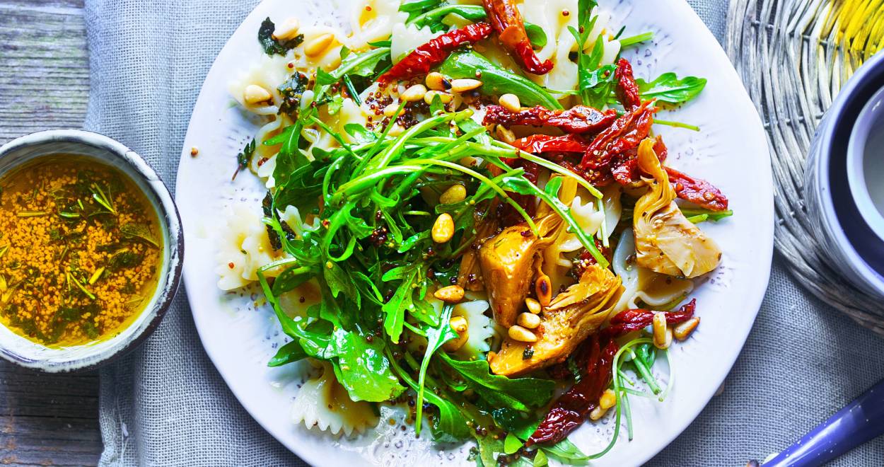 Mediterraner Nudelsalat auf einem Teller bestreut mit Pinienkernen, daneben eine Schale mit Vinaigrette