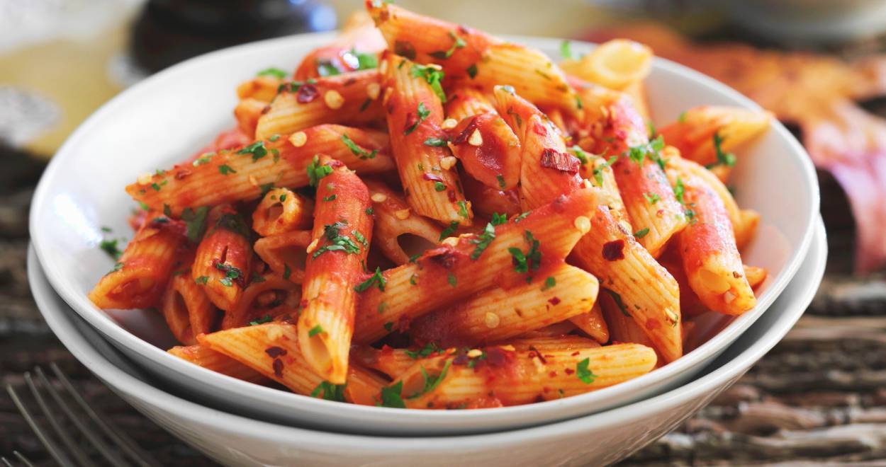 Penne all’arrabbiata in einem tiefen Teller auf einem Tisch, daneben eine Pfeffermühle
