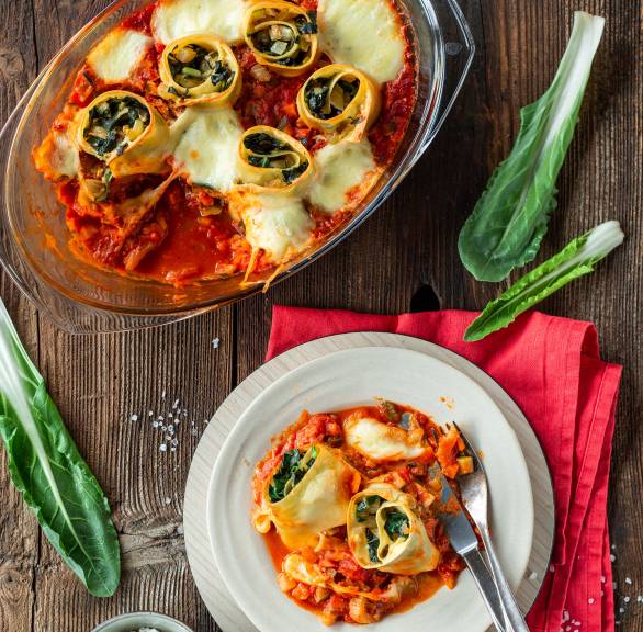 Zwei Mangold-Lasagne-Schnecken gefüllt mit Ricotta auf einem teller in Tomatensauce, daneben die Auflaufform