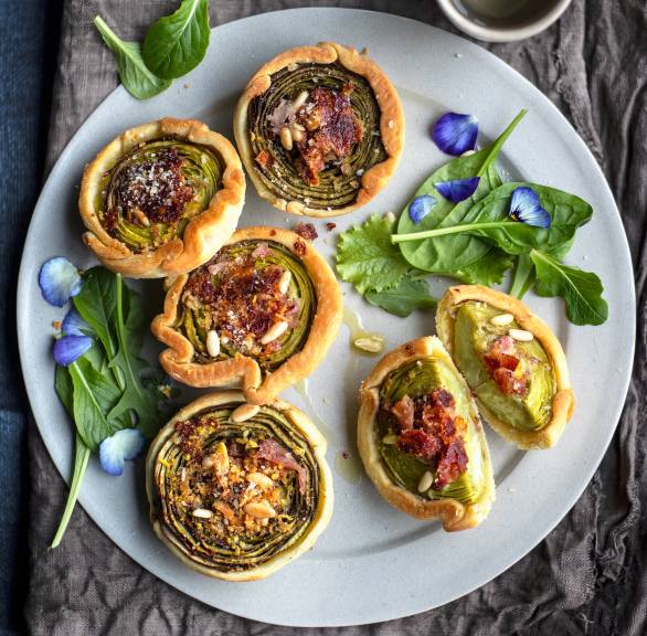 Mehrere selbstgebackene Artischocken-Törtchen aus Mürbeteig mit Parmaschinken und Käse getoppt von Pinienkernen auf einer Servierplatte