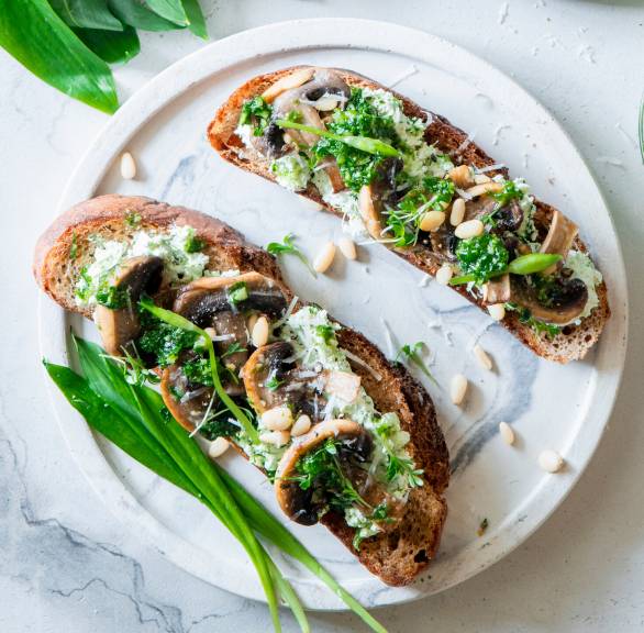 Bruschetta mit gebratenen Pilzen und Bärlauchpesto auf einem Teller, daneben frischer Bärlauch auf einem Tisch