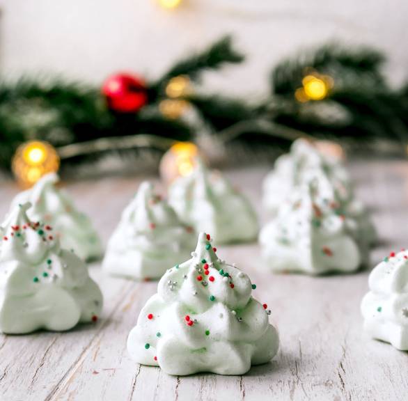 Tannenbäumchen aus Baiser auf einem Holztisch, dahinter leuchtende Weihnachtsdeko