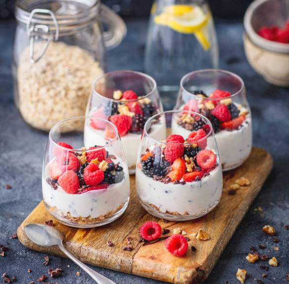 Schichtdessert in vier Gläsern getoppt mit Beeren, dahinter Granola und eine Karaffe mit Wasser auf einem Tisch