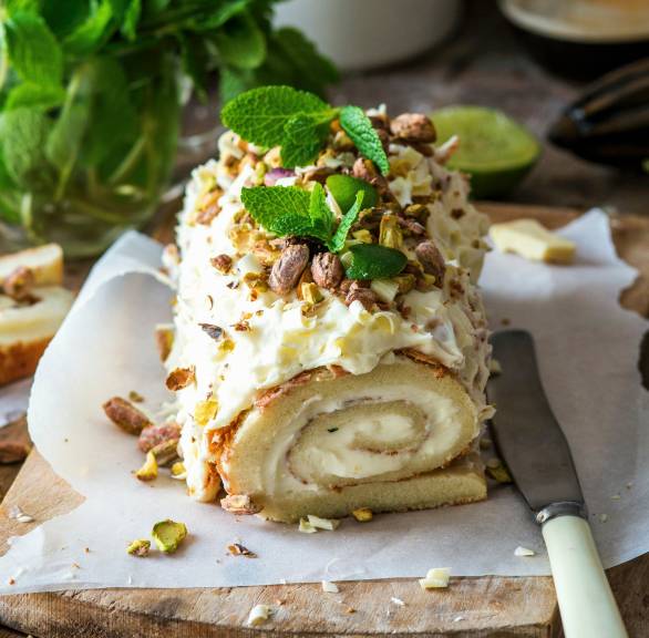 Pistazien-Biskuitrolle mit Mascarponecreme-Füllung angeschnitten auf einem Holzbrett garniert mit weißer Schokolade und Minzblättern