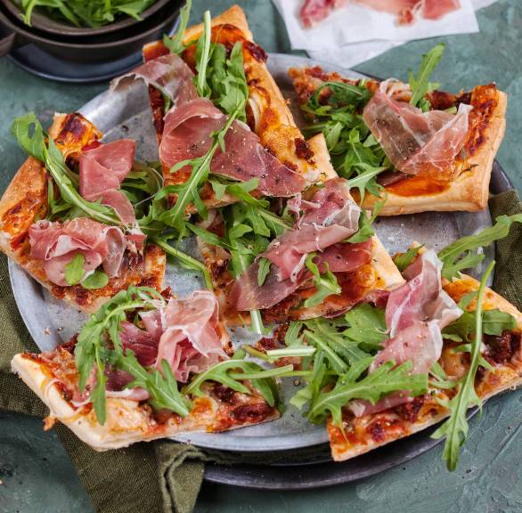 Mehrere Stücke Blätterteigpizza mit Parmaschinken, Mozzarella und Rucola angerichtet auf einem Teller