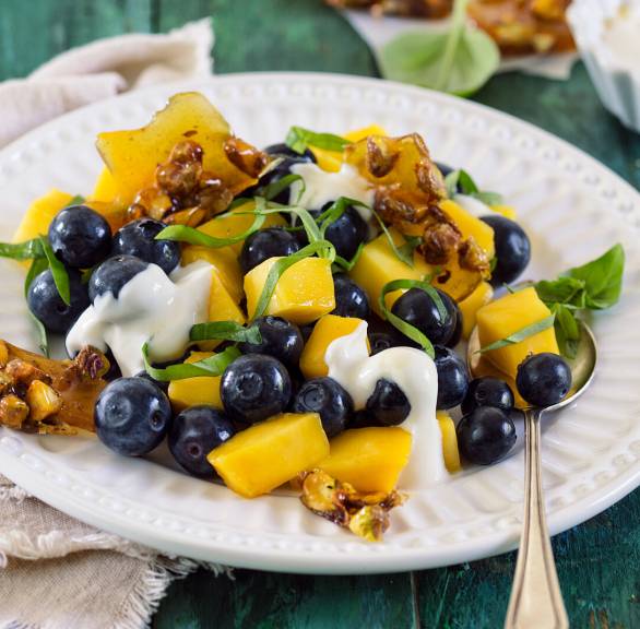Blaubeer-Mango-Salat auf einem Dessertteller getoppt von Joghurt-Mascarponecreme und Pistazien-Krokant