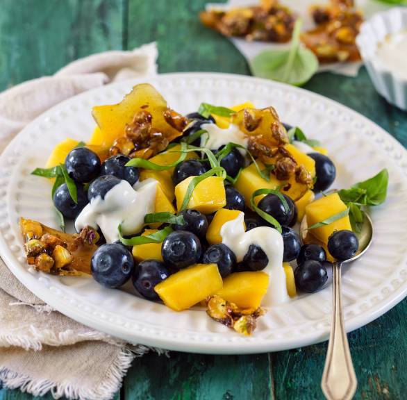 Blaubeer-Mango-Salat auf einem Dessertteller getoppt von Joghurt-Mascarponecreme und Pistazien-Krokant