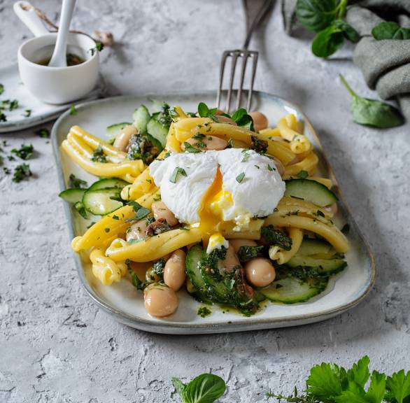 Nudelsalat aus Strozzapreti auf einem rechteckigen Teller, darauf ein pochiertes Ei, das gerade ausläuft