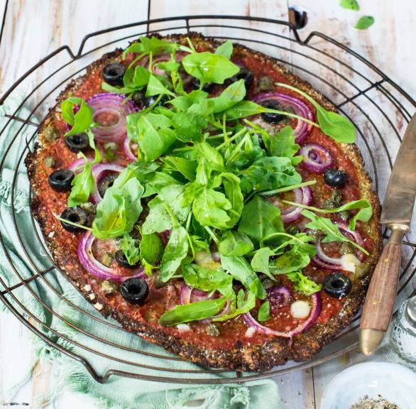 Ovale Low-Carb-Pizza mit Brokkoli-Käseboden und Belag aus Tomatensoße, Oliven, Kapern, Käse und Rucola auf einem Küchengitter
