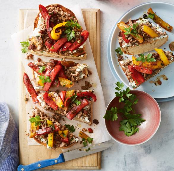 Großes Bruschetta in Streifen geschnitten auf einem Holzbrett getoppt mit Paprikasteifen, Hackfleisch und Sultaninen