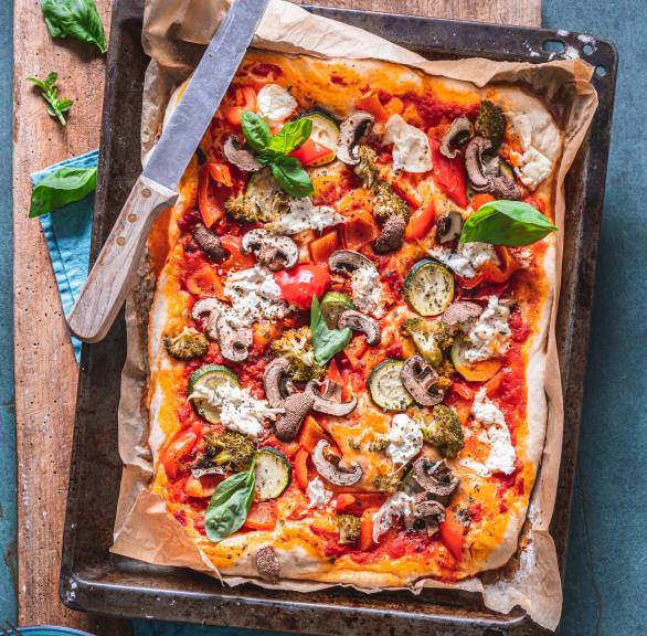 Pizza auf einem Blech belegt mit Champignons, Zucchini und Brokkoli auf einem Holzbrett