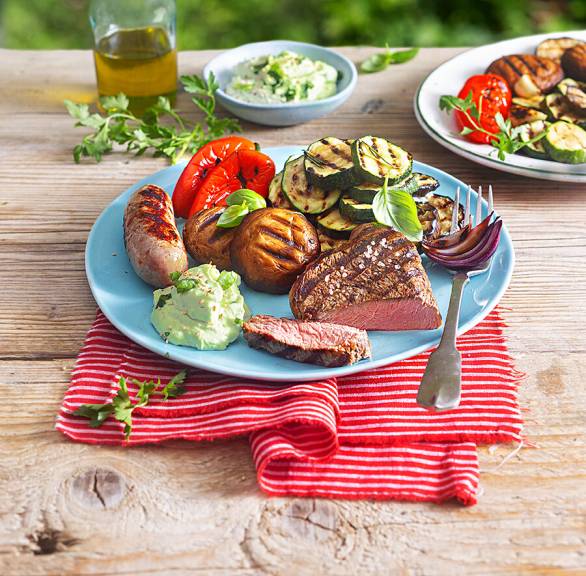 Mediterraner Grillteller mit Steak, Salsiccia, Gemüse, Avocado-Schmand-Dip und Knoblauch-Ciabatta auf einem Holztisch im Garten
