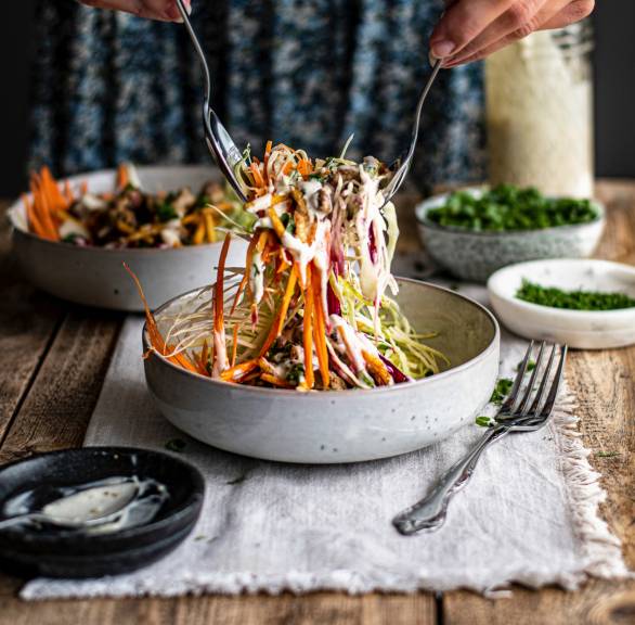 Krautsalat wird mit zwei Löffeln von einer Frau aus einer Schüssel gehoben, daneben Joghurt-Dressing in einem Glas