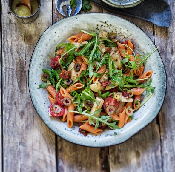 Salat aus roten Penne auf einem Teller mit Rucola und getrockneten Tomaten, daneben eine Schale mit grünen Oliven