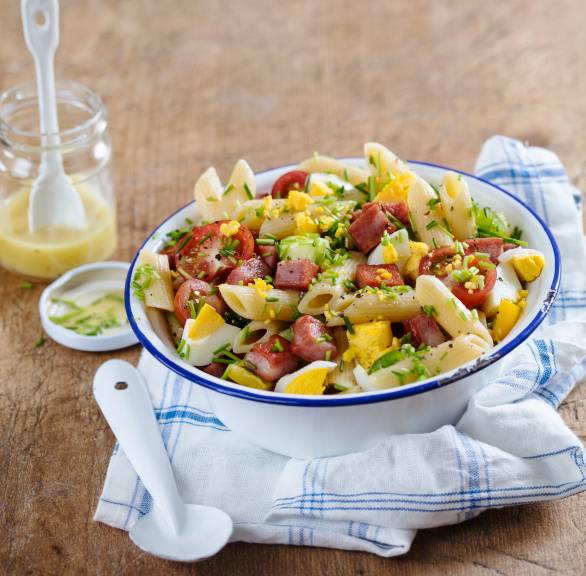 Penne-Salat in einer Schüssel getoppt mit Mortadella, Zucchini und Eierspalten, daneben Dressing in einem Glas