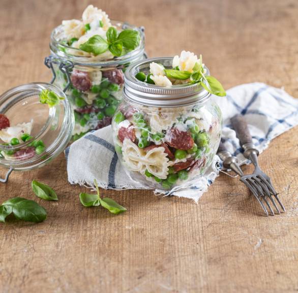 Farfalle-Salat geschichtet in zwei Weckgläser mit Cabanossischeiben und Joghurtdressing auf einem Tisch