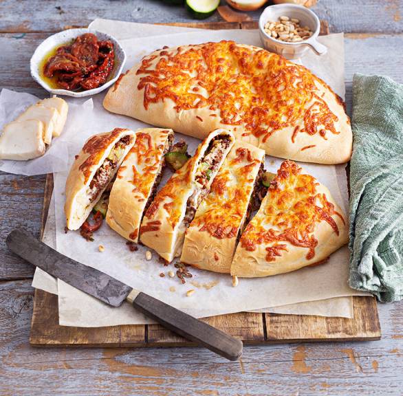 Zwei gebackene Calzones gefüllt mit Zucchini und Lammhackfleisch auf einem Holzbrett, davon eine in Streifen geschnitten