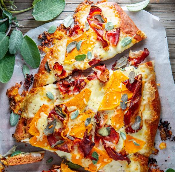 Dinkelpizza mit Butternutkürbis, Parmaschinken und Salbei in drei Teile geschnitten auf einem Tisch