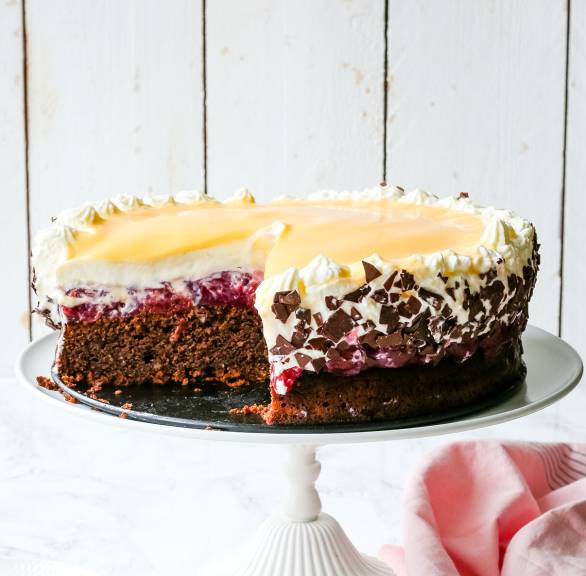Eierlikörtorte mit Kirschen angeschnitten auf einer Tortenplatte, davor ein Stück des Torte auf einem Teller