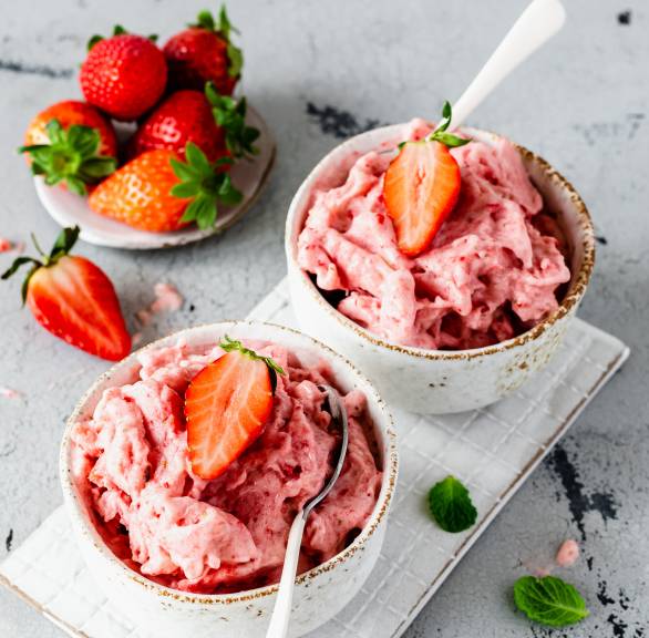 Zwei Portionen Erdbeer-Nicecream mit Banane in Schälchen auf einem Holztisch garniert mit frischen Erdbeeren