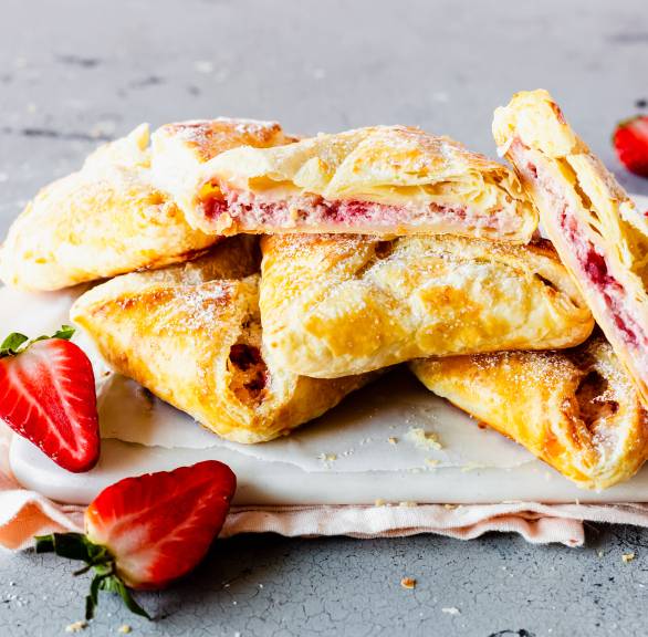 Fünf Blätterteigtaschen gefüllt mit Erdbeeren und Ricotta auf einem Teller, eine halbiert mit gut sichtbarer Füllung