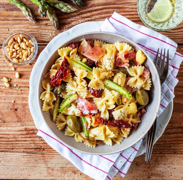 Nudelsalat mit Farfalle, grünem Spargel, Artischocken und Schinken angerichtet in einer Salatschüssel auf einem Holztisch
