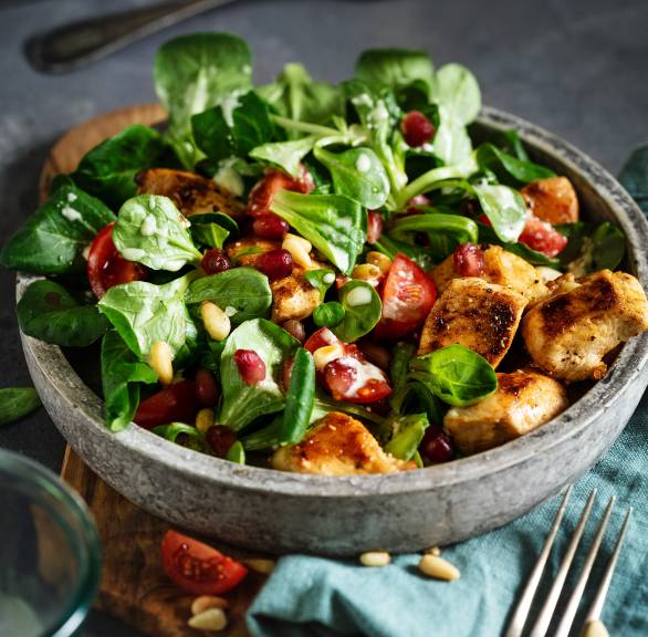 Feldsalat mit gebratenen Hähnchenstücken getoppt von Pinien- und Granatapfelkernen, daneben Parmesandressing