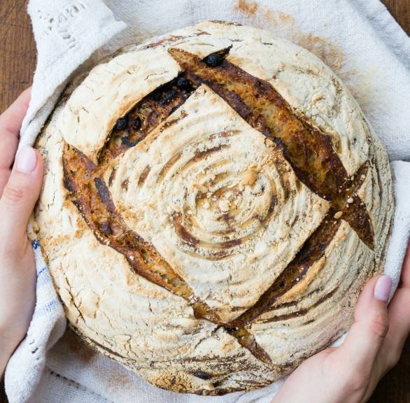 Rundes Rosinenbrot in den Händen einer Frau über einem Holztisch
