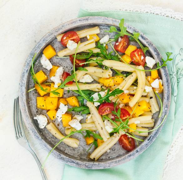 Pastasalat auf einem Teller mit Mangostücken, Schafskäse und Tomaten, daneben Besteck auf einem Tisch
