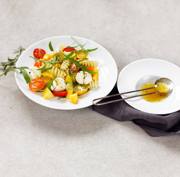 Pastasalat mit Tomaten und Mangostücken auf einem Teller, daneben Dressing in einer Schüssel