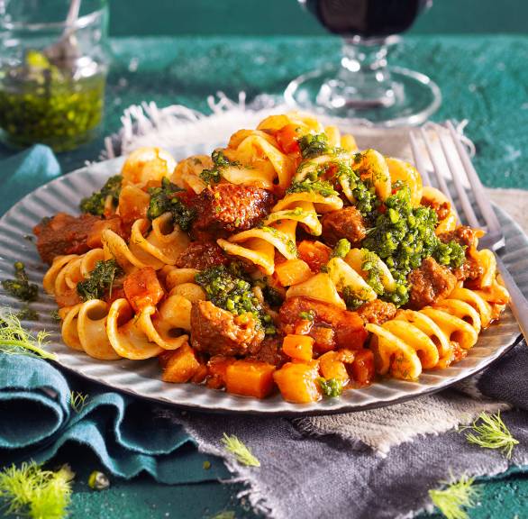 Kalbsragout mit Fusilli auf einem Teller getoppt von Fenchel-Pistazien-Pesto, daneben ein Glas Rotwein auf einem Tisch