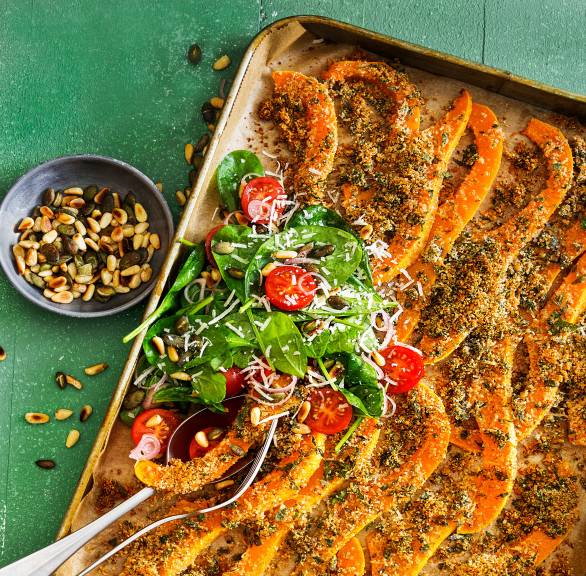 Gebackene Kürbisspalten mit Parmesankruste auf einem Backblech, daneben eine Schale Nüsse