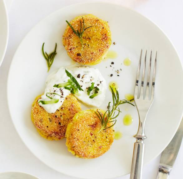 Mehrere gebratene Polenta-Bratlinge mit Schinken und Rosmarin angerichtet auf einem Teller mit Gurken-Minz-Dip