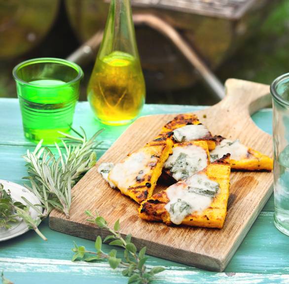 Gegrillte Polentaecken auf einem Holzbrett getoppt mit Gorgonzola, daneben eine Getränkekaraffe auf einem Tisch