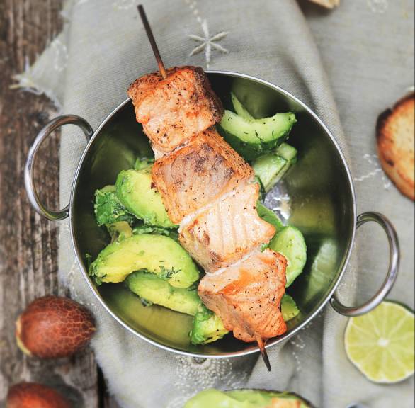 Gegrillter Lachs auf einem Spieß auf Gurkensalat, daneben Avocadodip und halbierte Avocados