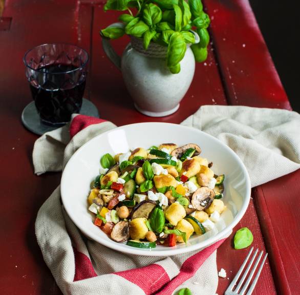 Gnocchi-Gemüse-Pfanne in einem tiefen Teller getoppt mit Feta auf einem Tisch, daneben frische Kräuter