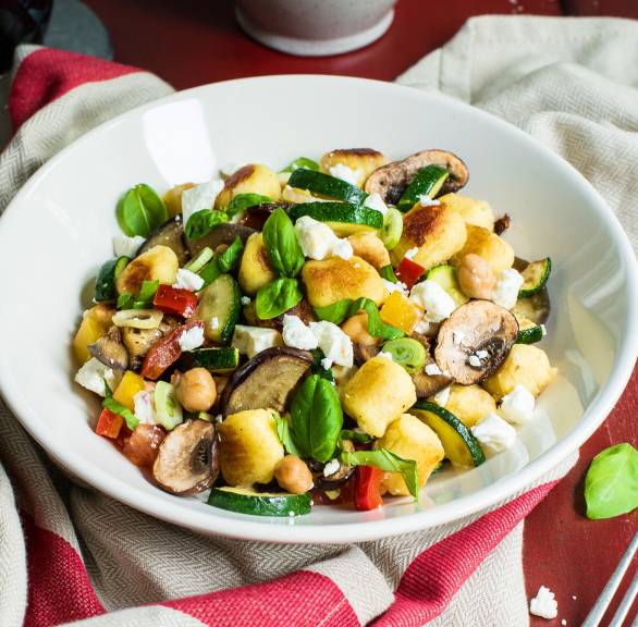 Gnocchi-Gemüse-Pfanne in einem tiefen Teller getoppt mit Feta auf einem Tisch, daneben frische Kräuter