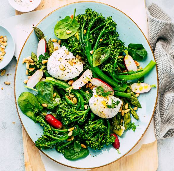 Gemüsesalat mit Spinat, Spargel und Brokkolini getoppt von Mozzarella, Dressing und Pinienkernen auf einem Teller