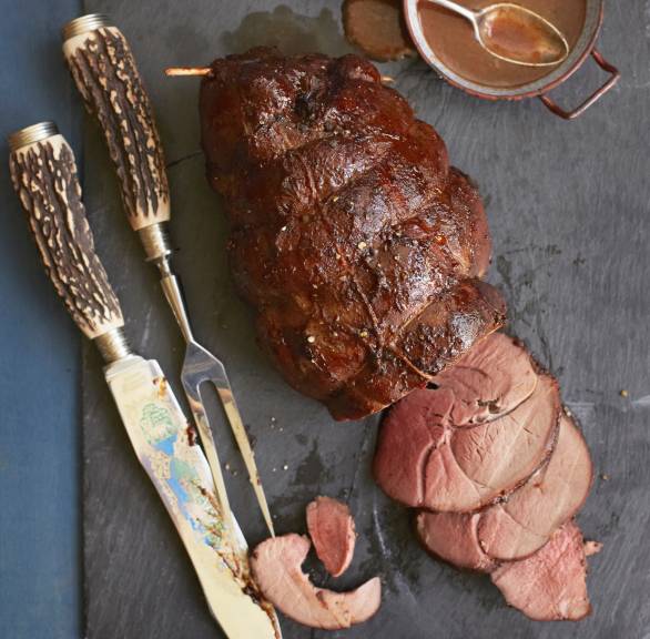 Geschmorte Rehkeule angeschnitten auf einer Servierplatte, daneben dunkle Soße in einer Sauciere
