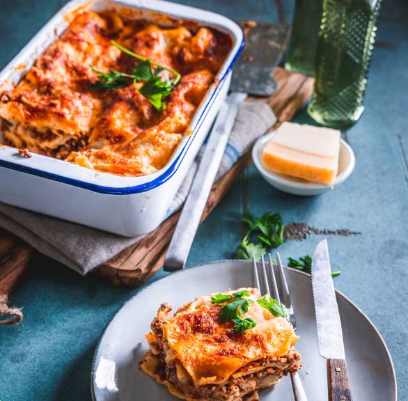 Glutenfreie Lasagne in einer Auflaufform, daneben ein Stück auf einem Teller und Parmesan