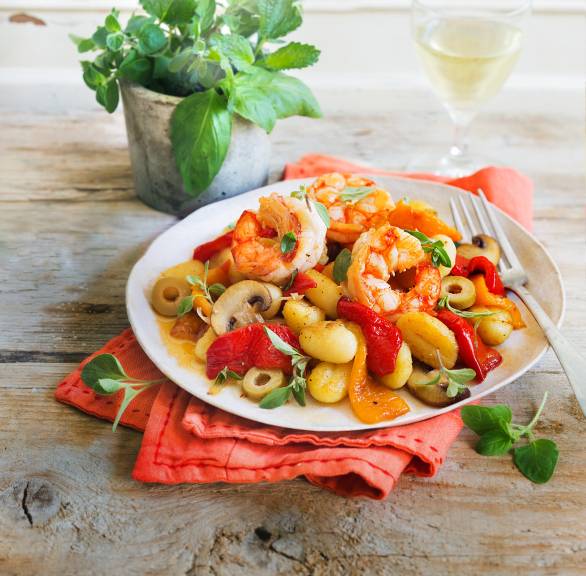 Gnocchisalat auf einem Teller getoppt von Antipasti und Garnelen, daneben ein Topf Basilikum auf einem Tisch
