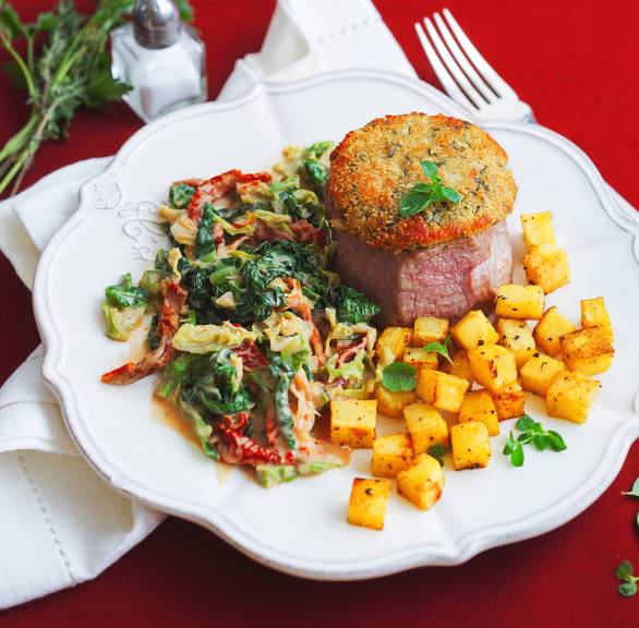 Gratiniertes Kalbsmedaillon mit Kräuterhaube auf einem Teller mit Kartoffelwürfeln und Wirsinggemüse in Sauce