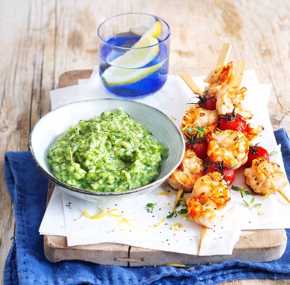 Spinatrisotto auf einem tiefen Teller angerichtet auf einem Holzbrett, daneben mit Garnelenspießen mit Tomaten
