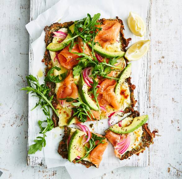 Rechteckige Low-Carb-Pizza mit Grünkohl-Käseboden und Räucherlachs-Avocado-Belag angeschnitten auf einem Holzbrett