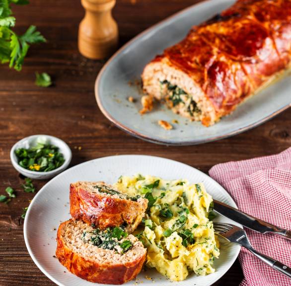 Ein Stück Hackbraten mit Mangold-Ricotta-Füllung, dahinter der restliche Hackbraten im Ganzen auf einem Holztisch