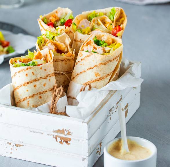 Hähnchen-Wraps nebeneinander in einer kleinen Holzkiste, daneben ein Schälchen mit Caesar-Salad-Creme