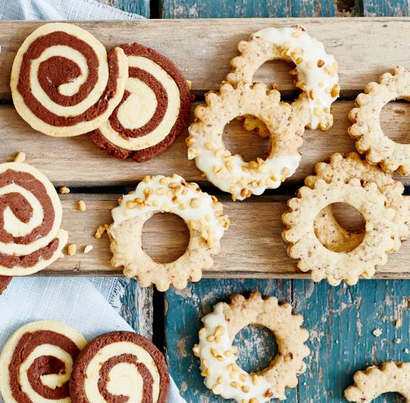 Selbst gebackene Haselnussplätzchen in Kringelforrm dekoriert mit Krokant und weißer Schokolade neben anderen Weihnachtsplätzchen auf einem Holzbrett